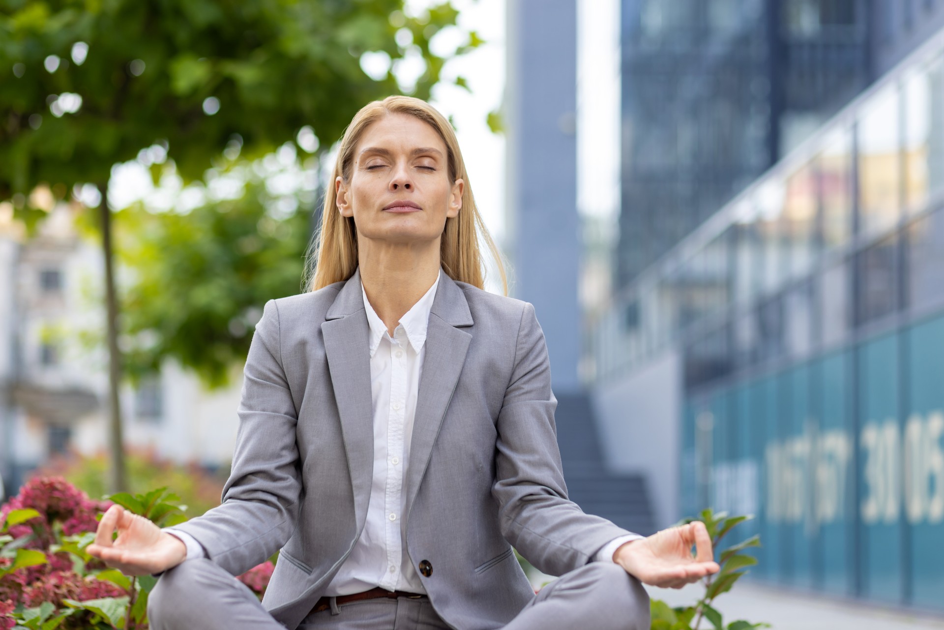 Tired young business woman sitting on bench outside office work building in lotus position with closed eyes, meditating and resting